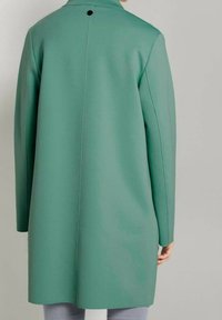 Muntgroene jas met een strak ontwerp, voorzien van een hoge kraag, lange mouwen en een enkele knoop aan de achterkant. Gladde textuur en eenvoudige lijnen.