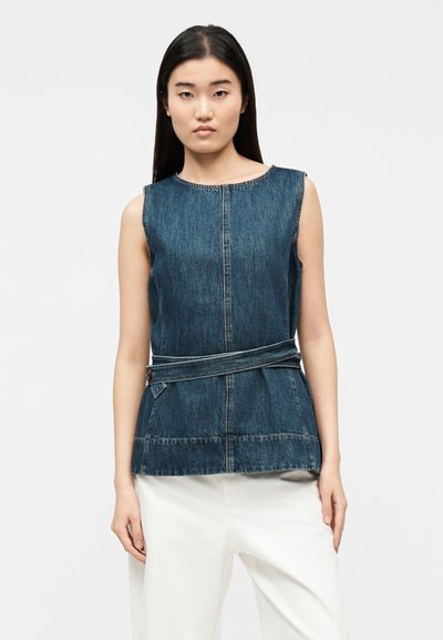 Femme aux cheveux longs et foncés portant un haut en jean sans manches avec une ceinture latérale et un pantalon blanc, se tenant devant un fond uni.