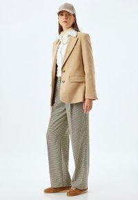 Beige säädetty bleiseri valkoisen nappipaidan päällä, yhdistettynä hounds-tooth-kuvioisiin housuihin ja ruskeisiin slip-on-kengiin, korostettuna ruutulakilla.