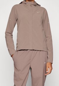 Chaqueta Nike ligera en color malva con capucha, mangas largas, puños elásticos y detalles reflectantes en las mangas. Textura de tela suave.