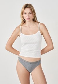 Mujer con cabello largo y rubio, vistiendo una blusa sin mangas blanca y ropa interior de algodón gris, de pie contra un fondo claro y uniforme.