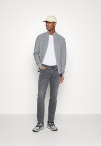 Gray zip-up sweater over a white t-shirt, paired with gray jeans and gray running shoes. The person wears a beige cap and stands against a white background.