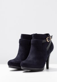 Botas altas de tacón en azul marino con una textura de ante suave, que cuentan con una correa decorativa negra y una hebilla plateada en el lateral.