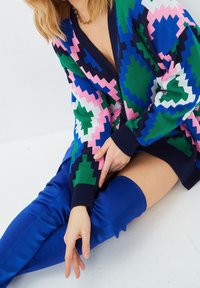 Colorful knitted cardigan featuring a geometric pattern in green, pink, and blue, with a V-neckline and ribbed cuffs. Paired with blue thigh-high boots.