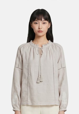 Donna con capelli neri lisci indossa una camicetta beige a maniche lunghe con scollo arricciato e laccetto con nappine, in piedi davanti a uno sfondo semplice.