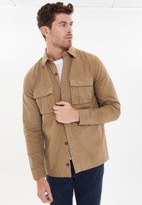 Beige Baumwollhemdjacke mit zwei Fronttaschen, Knopfverschluss und geradem Schnitt. Weißes Unterhemd ist unter der Jacke sichtbar.
