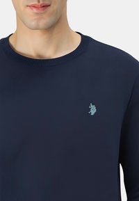 Maglione blu navy a coste con scollo rotondo e un piccolo logo ricamato azzurro chiaro sul petto. Texture liscia, maniche lunghe e taglio aderente.