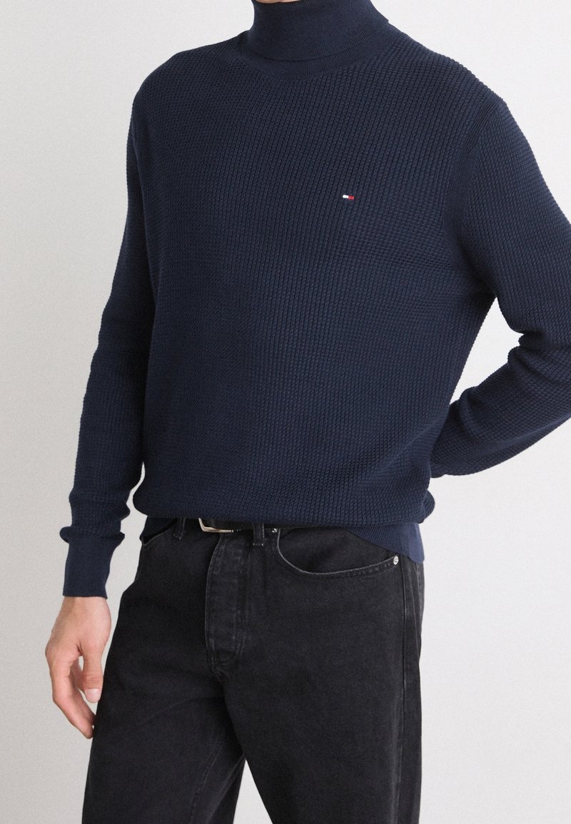 Homme portant un pull à col roulé texturé bleu marine et un jean noir, la main posée sur la hanche, sur un fond clair uni.