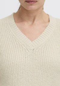 Lichtbeige gebreide trui met een V-hals, voorzien van een losse weefstructuur en geribde randen rond de halslijn.