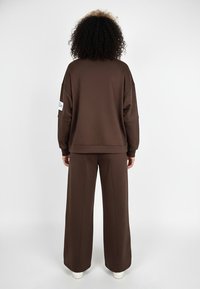 Brązowa bluza i pasujące szerokie spodnie wykonane z miękkiego materiału. Charakteryzują się luźnym krojem, opuszczonymi ramionami oraz detalem z etykietą na rękawie.