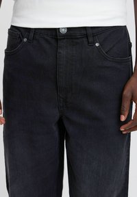Jeans de mezclilla negros con un diseño clásico de cinco bolsillos, con cierre de botón, remaches metálicos y una textura sutil.