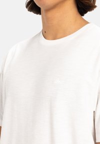 T-shirt bianca a maniche corte con collo rotondo, realizzata in un tessuto testurizzato e dotata di un logo ricamato discreto sul petto. Design semplice e minimalista.