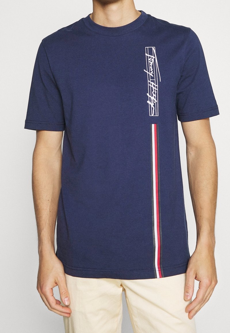 Námorně modré bavlněné tričko s krátkým rukávem. Obsahuje bílý grafický potisk na levé straně a vertikální akcent z tri-barvy pruhů.