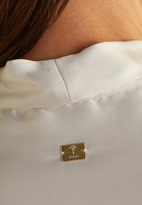 Collo in tessuto bianco con una superficie liscia, dotato di una piccola piastrina dorata sul retro, che mostra un design ad albero e il nome del marchio.