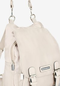 Mochila de tela beige con bolsillos con cremallera, cierre de solapa, correas ajustables y detalles de metal en color plata. Etiqueta de la marca visible.