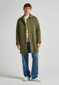 Olivengroen, lichtgewicht jas met knoopsluiting, zijzakken en een getailde kraag, gedragen over een roomkleurige trui met rode strepen en blauwe jeans.