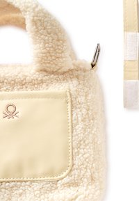 Beżowa torebka z futerka ekologicznego o miękkiej, teksturowanej powierzchni, wyposażona w gładką skórzaną kieszeń oraz charakterystyczne oznaczenie, uzupełniona odpinanym paskiem.