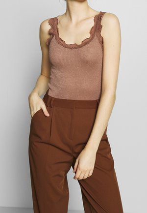 Femme portant un débardeur marron côtelé orné de dentelle et un pantalon marron taille haute, une main dans la poche, debout devant un fond uni.