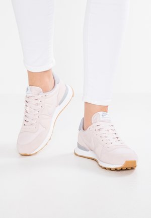 Osoba nosząca jasnoróżowe trampki Nike z białymi podeszwami i gumowymi podeszwami na zewnątrz, w połączeniu z białymi dopasowanymi spodniami na białej podłodze.