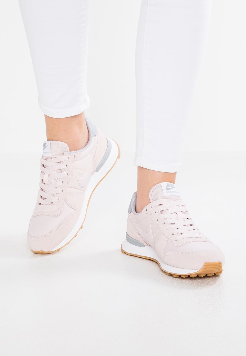 Personne portant des baskets Nike rose clair avec semelles blanches et semelles extérieures en gomme, associées à un pantalon blanc ajusté sur un sol blanc.