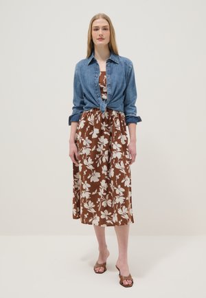 Femme portant une robe midi marron à fleurs avec une chemise en jean bleue nouée par-dessus et des sandales marron à talons ouvertes, debout devant un fond uni.