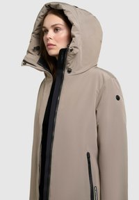 Beige hættejakke lavet af glat stof, med en sort lynlås, sidelommer og justerbar hætte med snore.