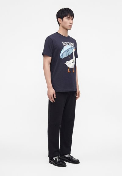 Άνδρας με μαύρο μπλουζάκι Moschino με γραφικό μιας χήνας που κρατάει μια μπλε ομπρέλα, μαύρο παντελόνι με φερμουάρ και μαύρα γυαλιστερά loafers.
