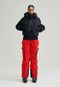 Jachetă izolată neagră cu două buzunare frontale cu fermoar, asortată cu pantaloni roșii cu fermoare laterale și accente negre. Construită pentru utilizare în aer liber.