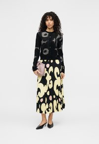 Cardigan negru cu modele circulare, asortat cu o fustă plisată galbenă, decorată cu desene abstracte negre și roz. Ține în mână o geantă mică roz.