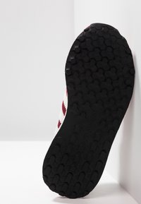 Svart gummisko sula med ett hexagonmönster, lutad mot en ljus yta. Har en vit överdel med röda zigzag-accenter.