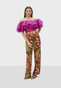 Lilla off-shoulder bluse med flæser, parret med brune mønstrede højtaljede bukser med farverige bladmotiver. Flade sandaler.