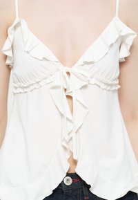 Haut blanc sans manches avec bords à volants, fermeture par nouage à l'avant et ourlet festonné, porté avec un jean en denim foncé.