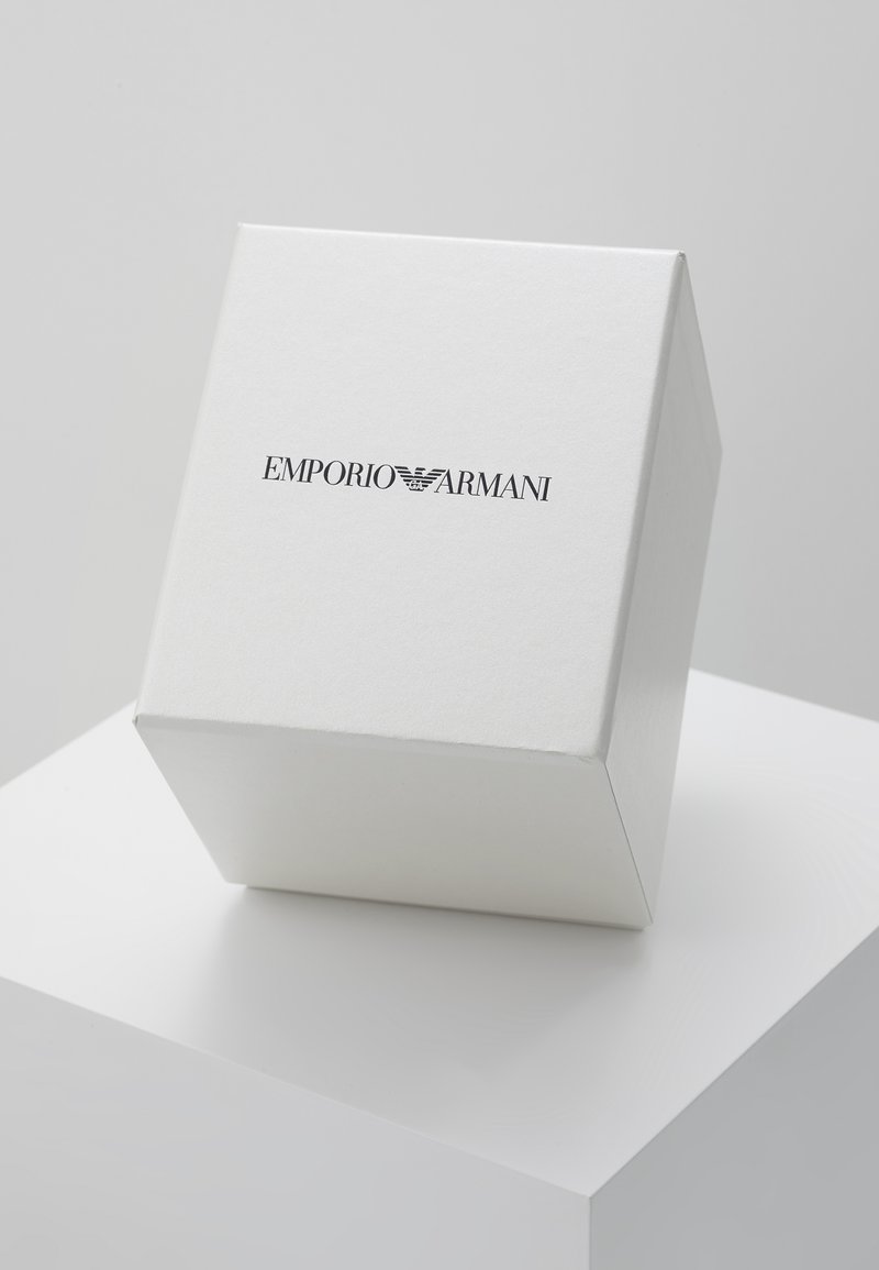 Weißer quadratischer Kasten aus strukturiertem Material mit dem schwarzen Logo "EMPORIO ARMANI", das auf einer weißen Präsentationsfläche platziert ist.