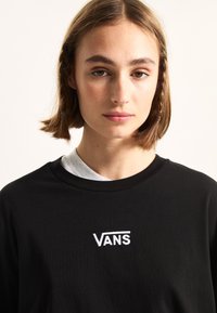 Svart bomullströja med rund halsringning, med vitt broderat "VANS"-logotyp på bröstet, lagrad över en grå skjorta.