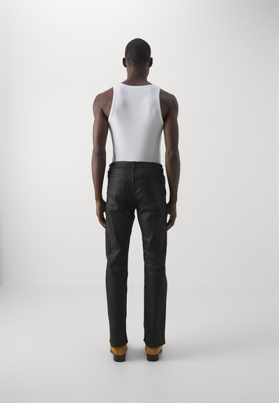 Schwarze Jeans mit geradem Schnitt, kombiniert mit einem schmal geschnittenen weißen Tanktop. Die Person trägt gelbe Stiefel. Rückansicht.