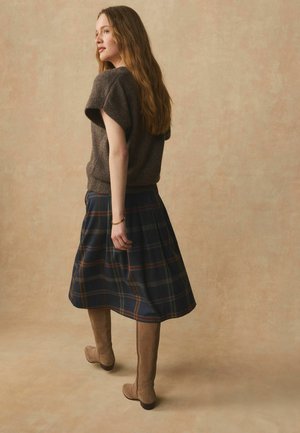 Kvinde med langt hår iført en brun kortærmet sweater, tartan nederdel i midi-længde, lysebrune knæhøje støvler og et guldbånd, stående foran en beige baggrund.