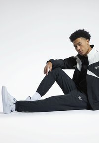 Schwarz-weiße sportliche Jacke mit Reißverschluss, schwarze Hose und weiße Sneakers. Das Outfit hat elastische Bündchen und ein kleines Logo auf der Jacke.