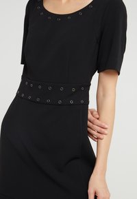 Vestito nero con scollo rotondo e maniche corte. Presenta una vita texturizzata adornata con occhielli in argento per un tocco di dettaglio in più.