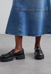 Mocassini in pelle nera con suola spessa e rigata e dettagli in oro. Abbinati a una gonna di jeans blu, che mette in mostra un look casual.
