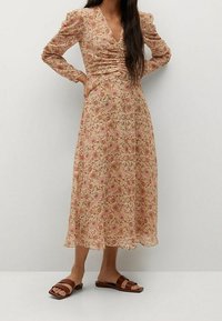 Femme portant une robe midi florale à manches longues avec un bustier froncé et des sandales plates marron, se tenant contre un mur clair et uni.