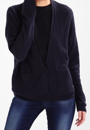 Personne portant un cardigan bleu marine foncé sur une chemise noire et un jean bleu, avec une main touchant leur cou.