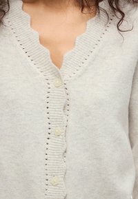 Cardigã cinza claro com decote ondulado, cinco botões e costura aberta decorativa nas bordas. Tecido de malha macio e texturizado.