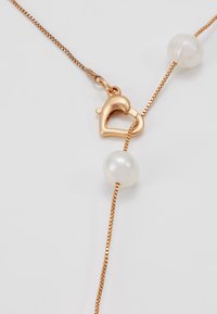 Collar de cadena en tono dorado que presenta un cierre en forma de corazón y dos perlas blancas. La cadena es delicada con una textura fina.