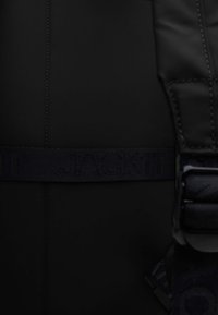 Mochila acolchada negra con una superficie texturizada, costuras prominentes y una asa tejida con la inscripción "JACKIT". Detalles oscuros y de hardware visibles.
