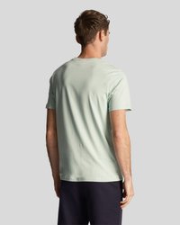 Homme portant un t-shirt à manches courtes vert clair et un short sombre, se tenant de dos contre un fond gris clair uni.