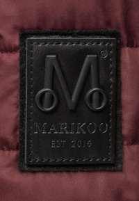 Černý kožený štítek s embosovaným logem "M" a textem "MARIKOO EST 2016", připevněný na bordové látce. Štítek má texturovaný povrch.