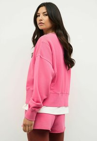 Felpa rosa oversize realizzata in misto cotone, con polsini e orlo a costine. Abbinata a pantaloni corti rosa coordinati, vista di lato.
