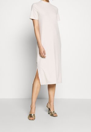 Mujer vestida con un vestido acanalado de manga corta y largo hasta la rodilla de color claro con una abertura lateral, combinado con sandalias de tacón alto abiertas de color dorado.
