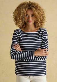 Námornícky a biely pruhovaný top s dlhým rukávom a lodičkovým výstrihom, so značkovou nášivkou na ramene, ktorý je skombinovaný so svetlými nohavicami.