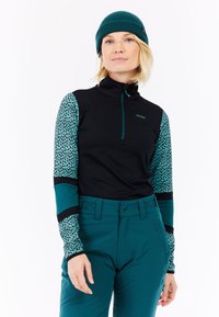 Zwarte rits-top met teal patroon mouwen, gecombineerd met teal broek. Geribbeld teal beanie. Opvallende patronen op de mouwen en contrasterende kleuren.